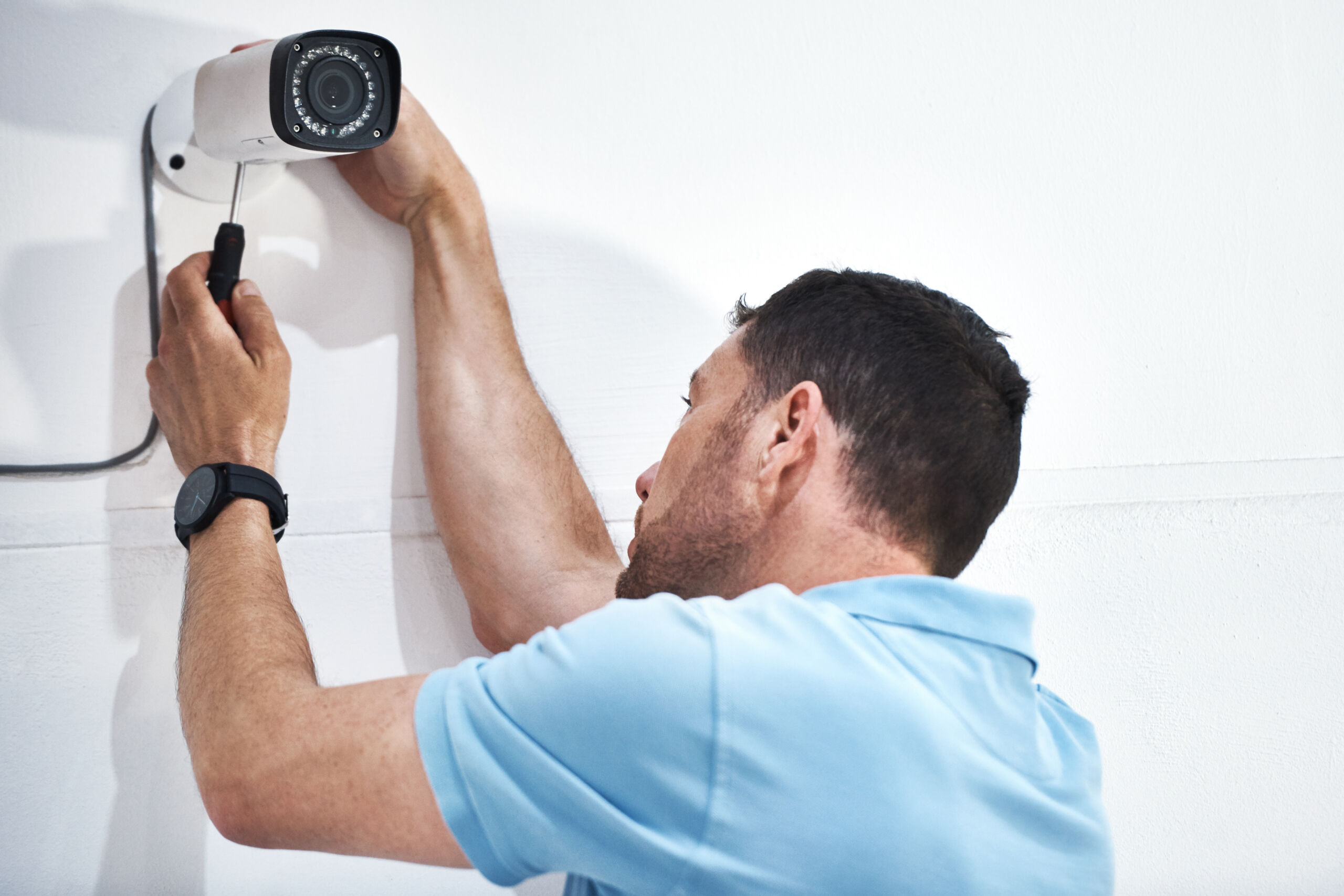 A man in a light blue shirt is installing a security camera on a white wall using a screwdriver.