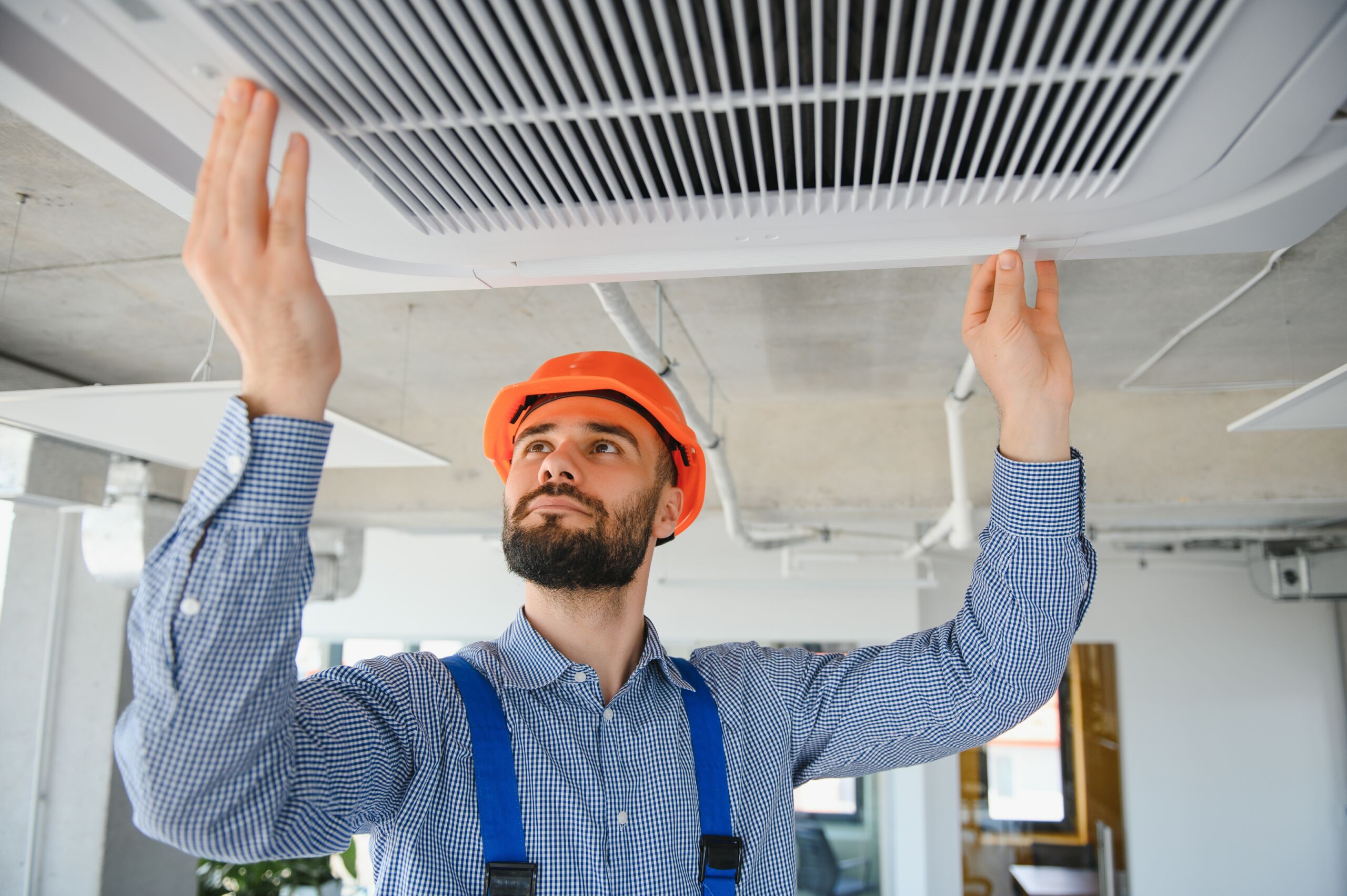 A man in a hard hat and safety braces inspects or repairs a ceiling air conditioning vent in an office space.