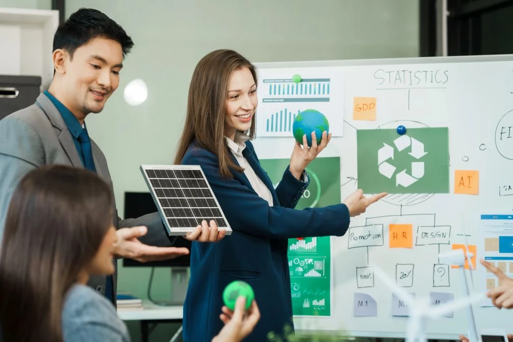 Three people in business attire present a solar panel and green props in front of a whiteboard with charts and a recycling symbol during a meeting on sustainability.