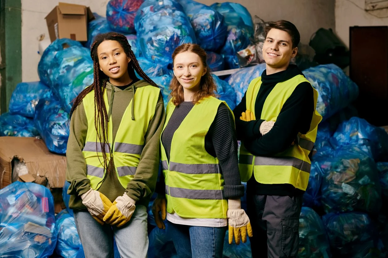 Three people in reflective vests and gloves stand in front of large piles of blue plastic bags, posing in a recycling or waste management facility.