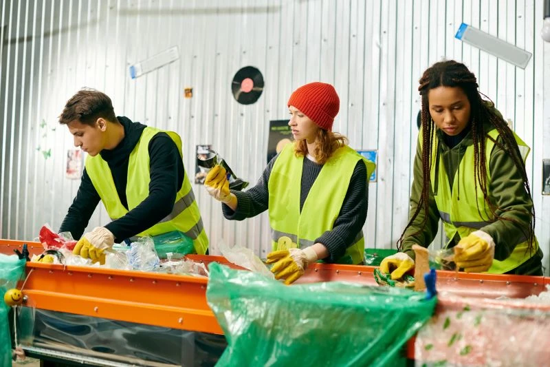 Three people wearing yellow safety vests and gloves sort recyclable materials on a conveyor belt in an industrial facility.