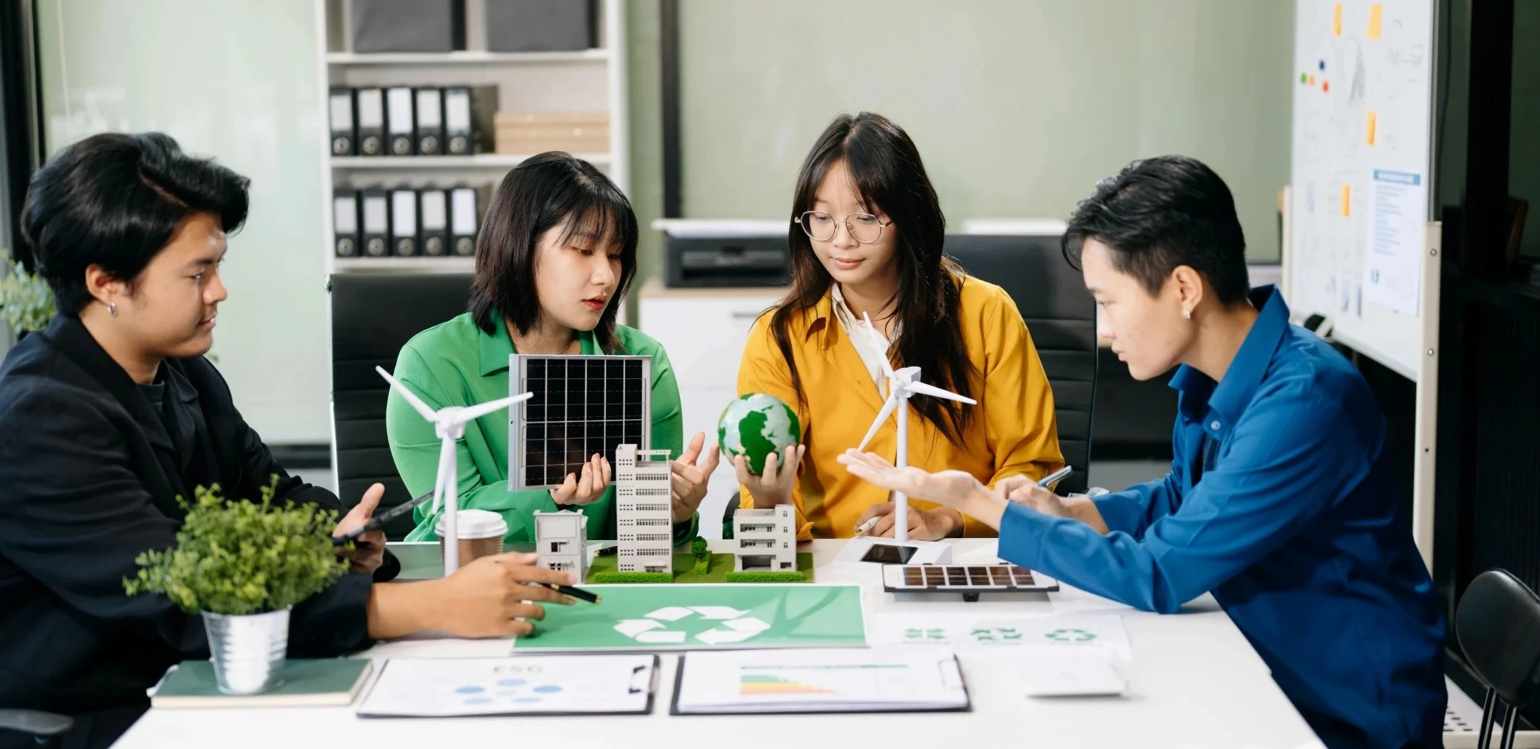 Four people at a table discuss renewable energy models, including wind turbines and solar panels, with documents, a globe, and a recycling symbol visible.