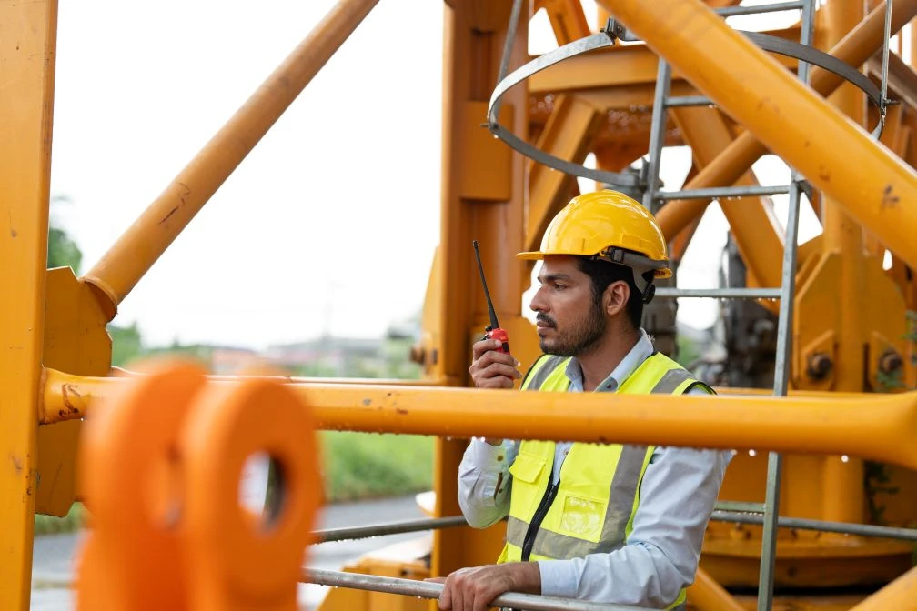 A construction worker in a yellow safety vest and hard hat uses a walkie-talkie whilst standing on a metal structure at a construction site.