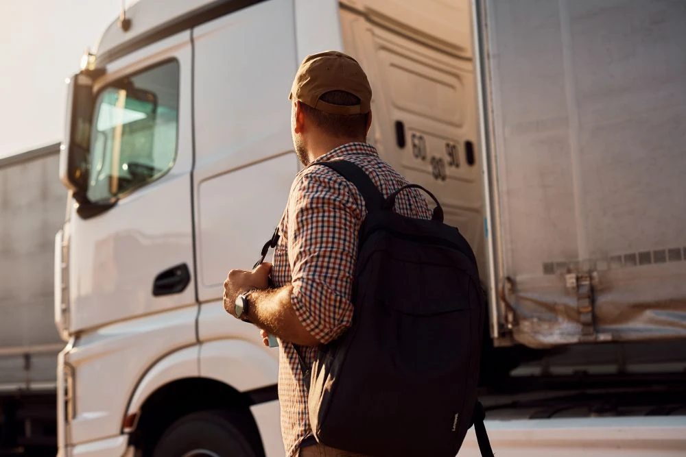 A man wearing a cap and rucksack stands beside a white lorry, facing away from the camera in daylight.