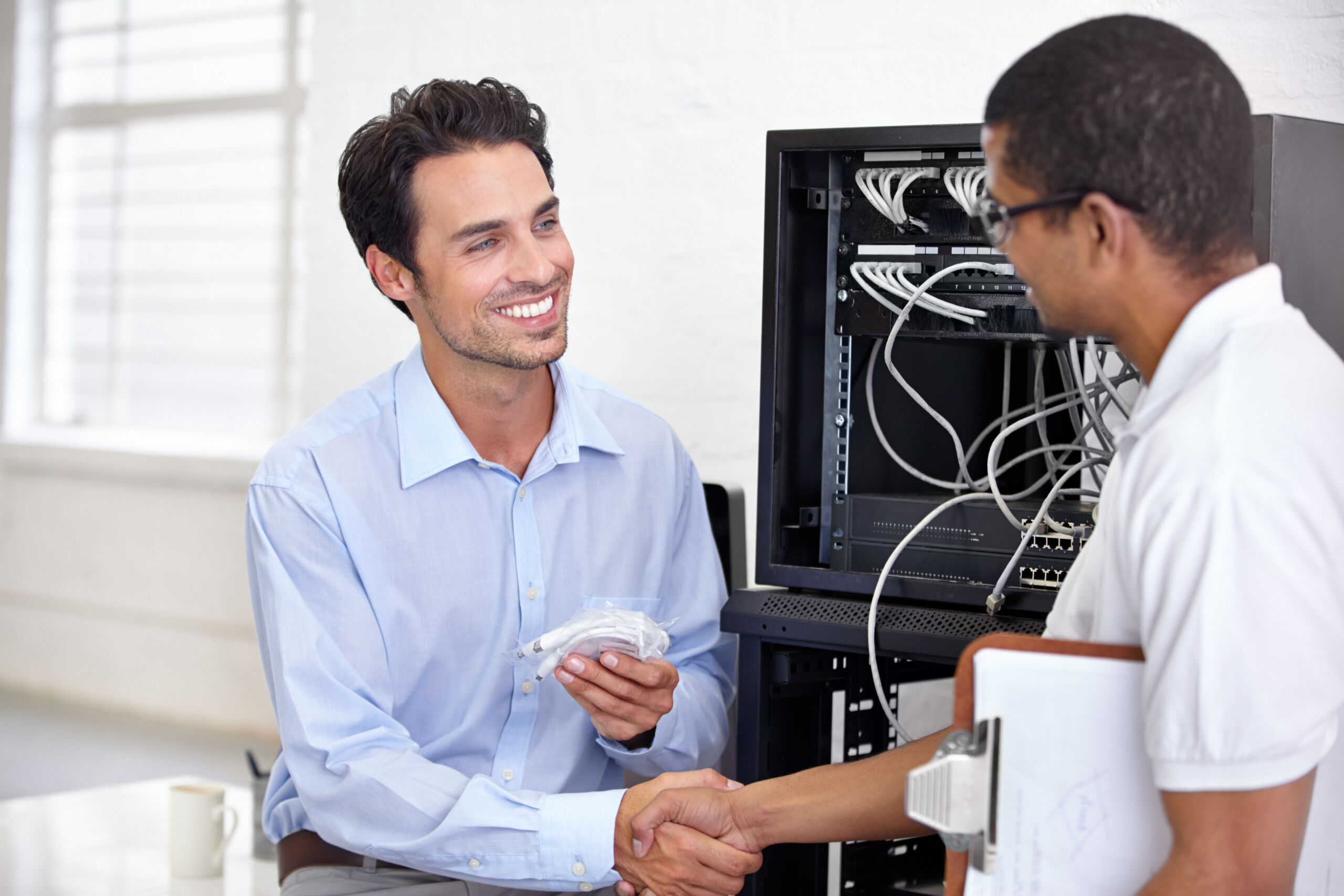 Two men shake hands in front of a server rack; one holds cables and smiles, and the other holds a clipboard.