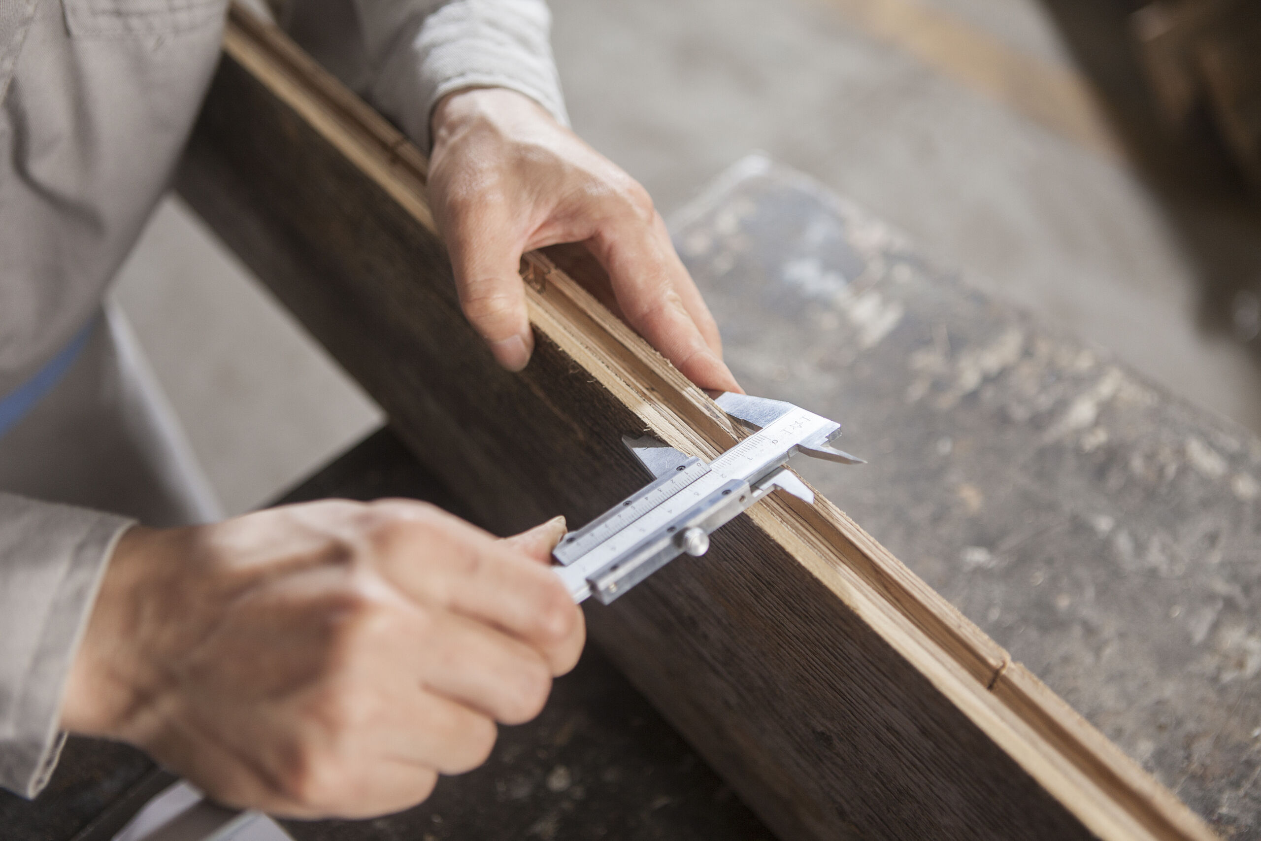 A person measures the thickness of a wooden plank with a vernier calliper on a workbench.