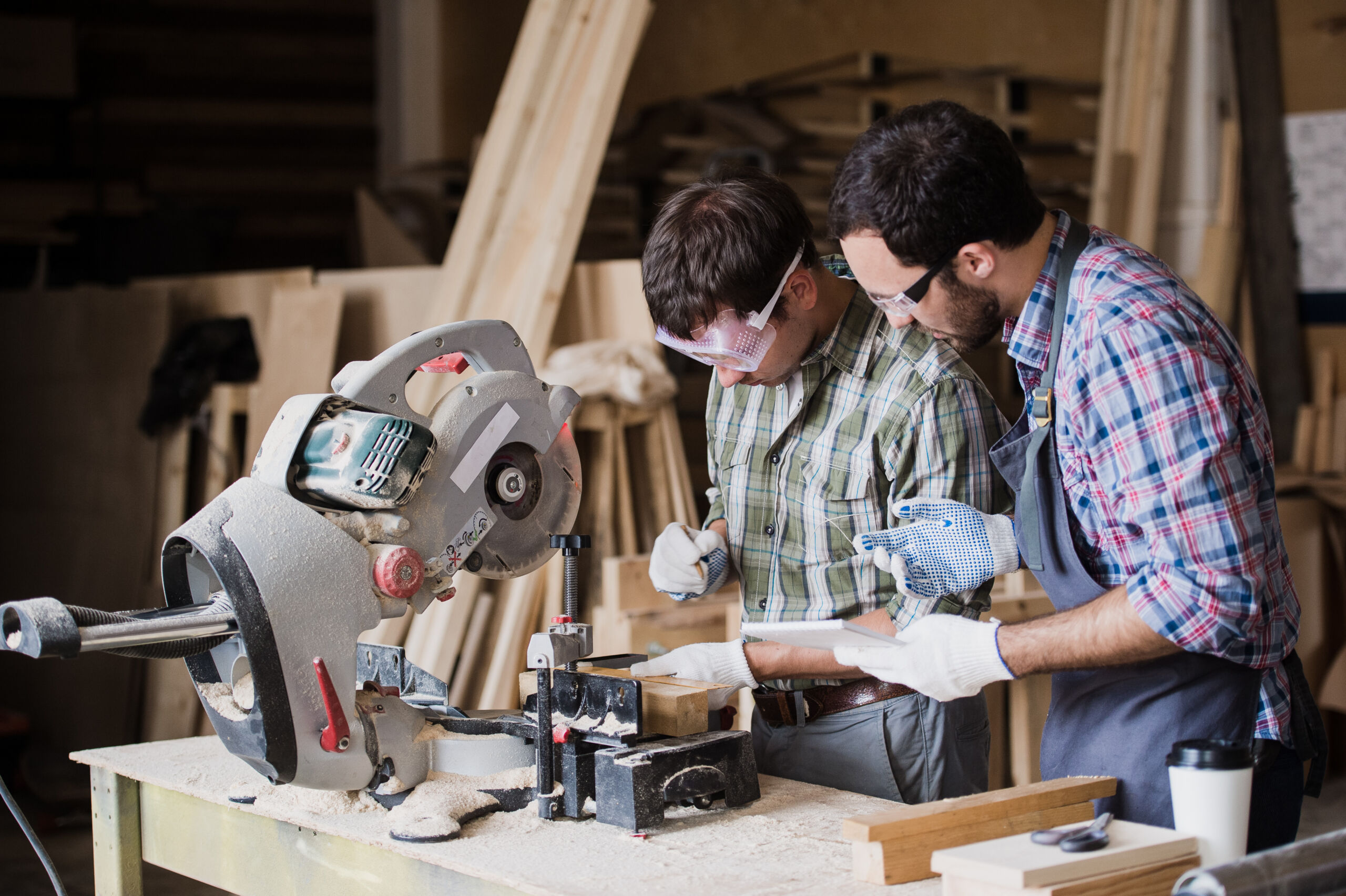 Two men in safety goggles and gloves operate a circular saw and work with timber in a carpentry workshop.