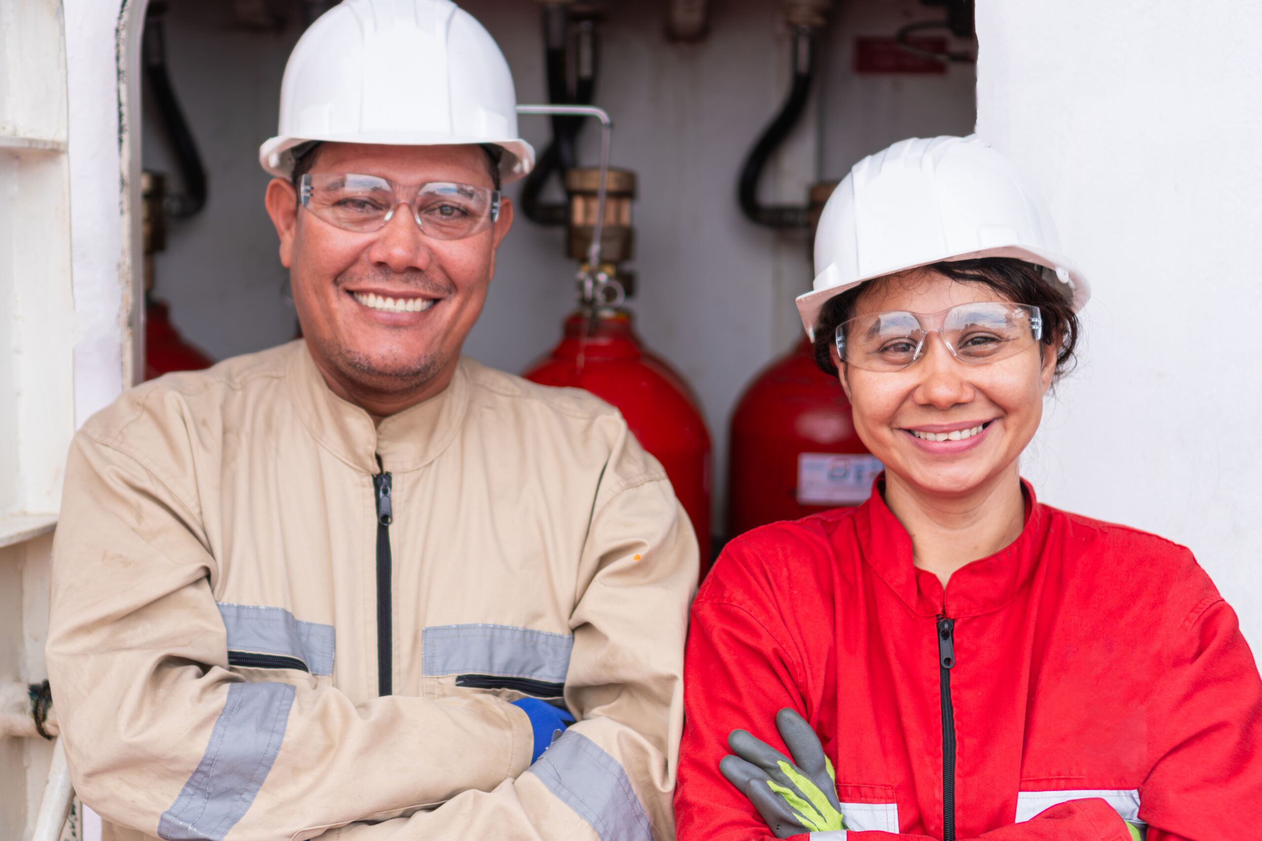 Two workers wearing hard hats, safety glasses, and overalls stand side by side, smiling, with red gas cylinders visible in the background.