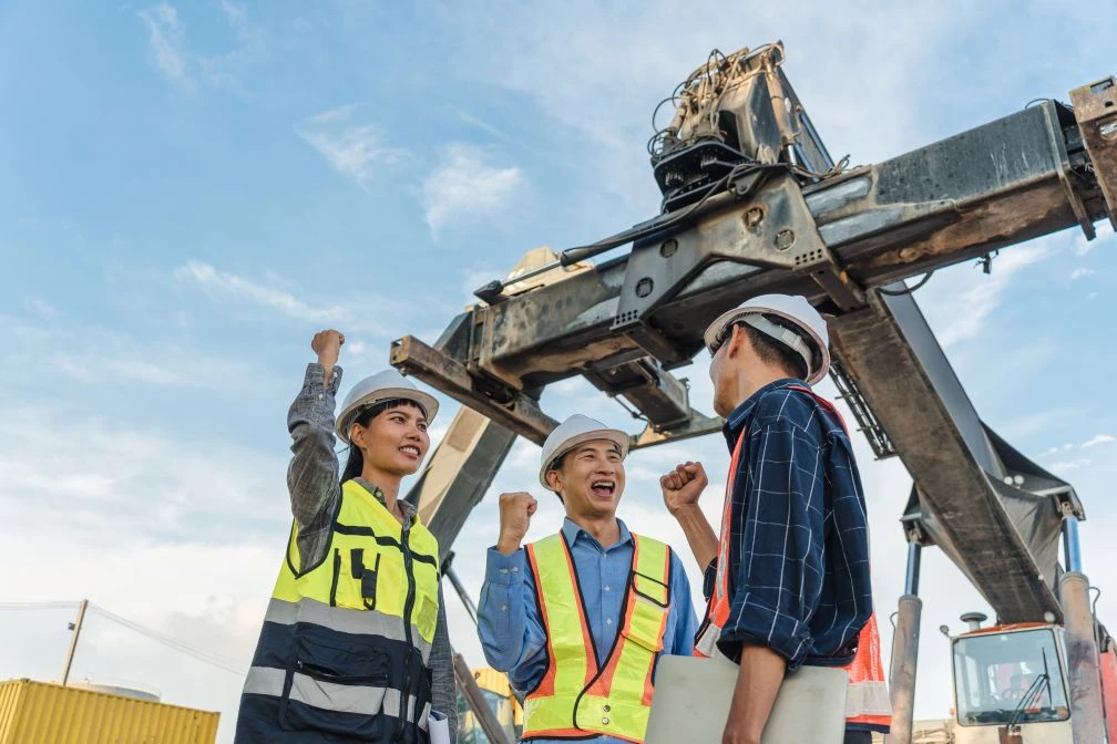 Three construction workers wearing safety gear stand under heavy machinery, raising their fists and smiling, with blue sky in the background.