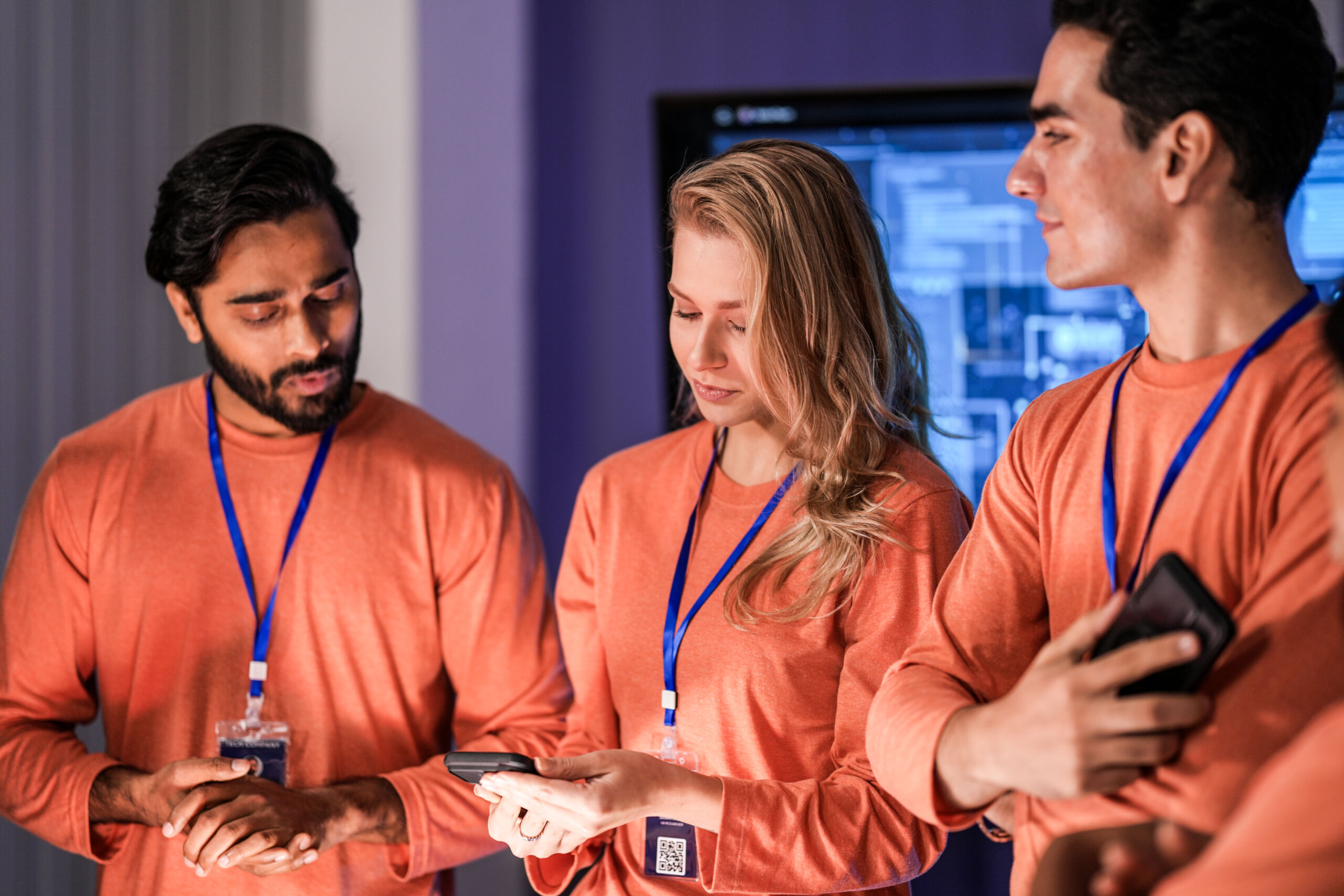 Three people wearing orange shirts and blue lanyards stand together indoors, looking at a mobile phone, with a screen displaying diagrams in the background.
