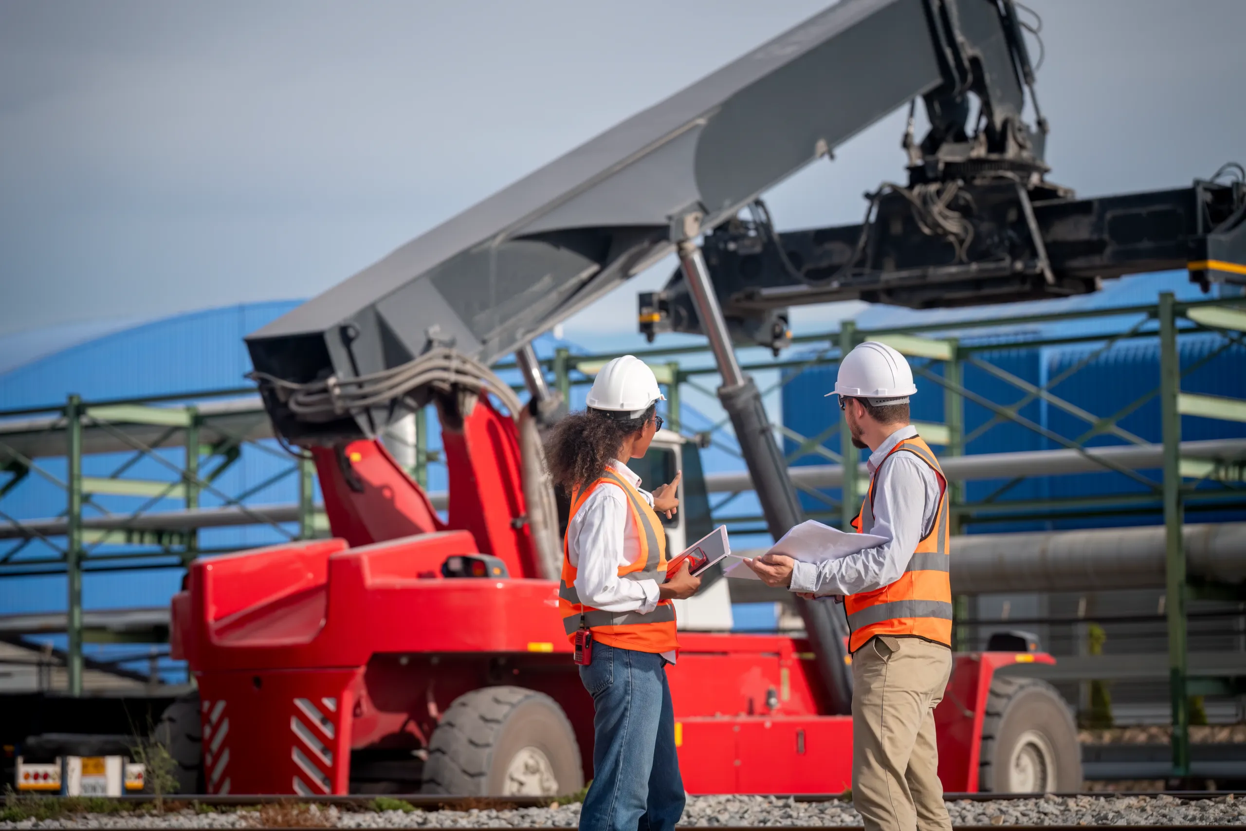 Two construction workers wearing safety vests and helmets discuss plans near a large red forklift at an industrial site.