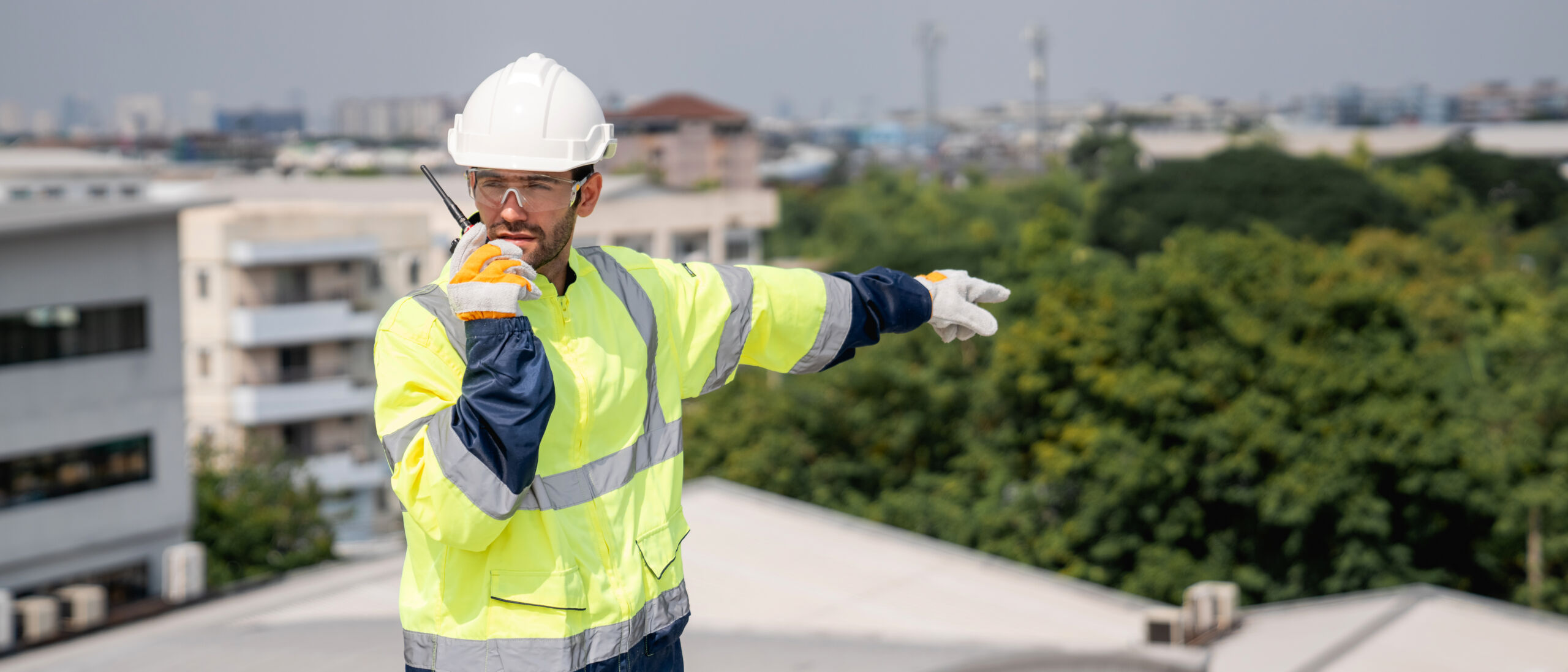 A construction worker in a hi-vis jacket and hard hat speaks into a radio while pointing, with buildings and trees visible in the background.