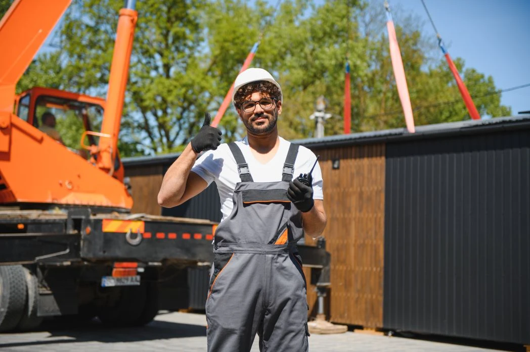 A construction worker in a white helmet and grey overalls stands outdoors near an orange crane lorry, giving a thumbs-up gesture with his right hand.