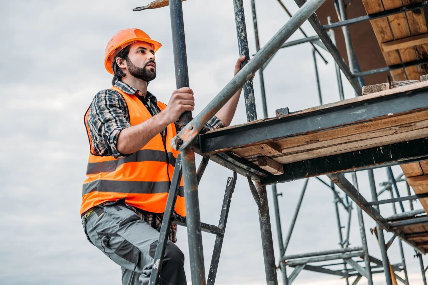 A construction worker in an orange safety vest and helmet climbs a metal scaffold at a building site.