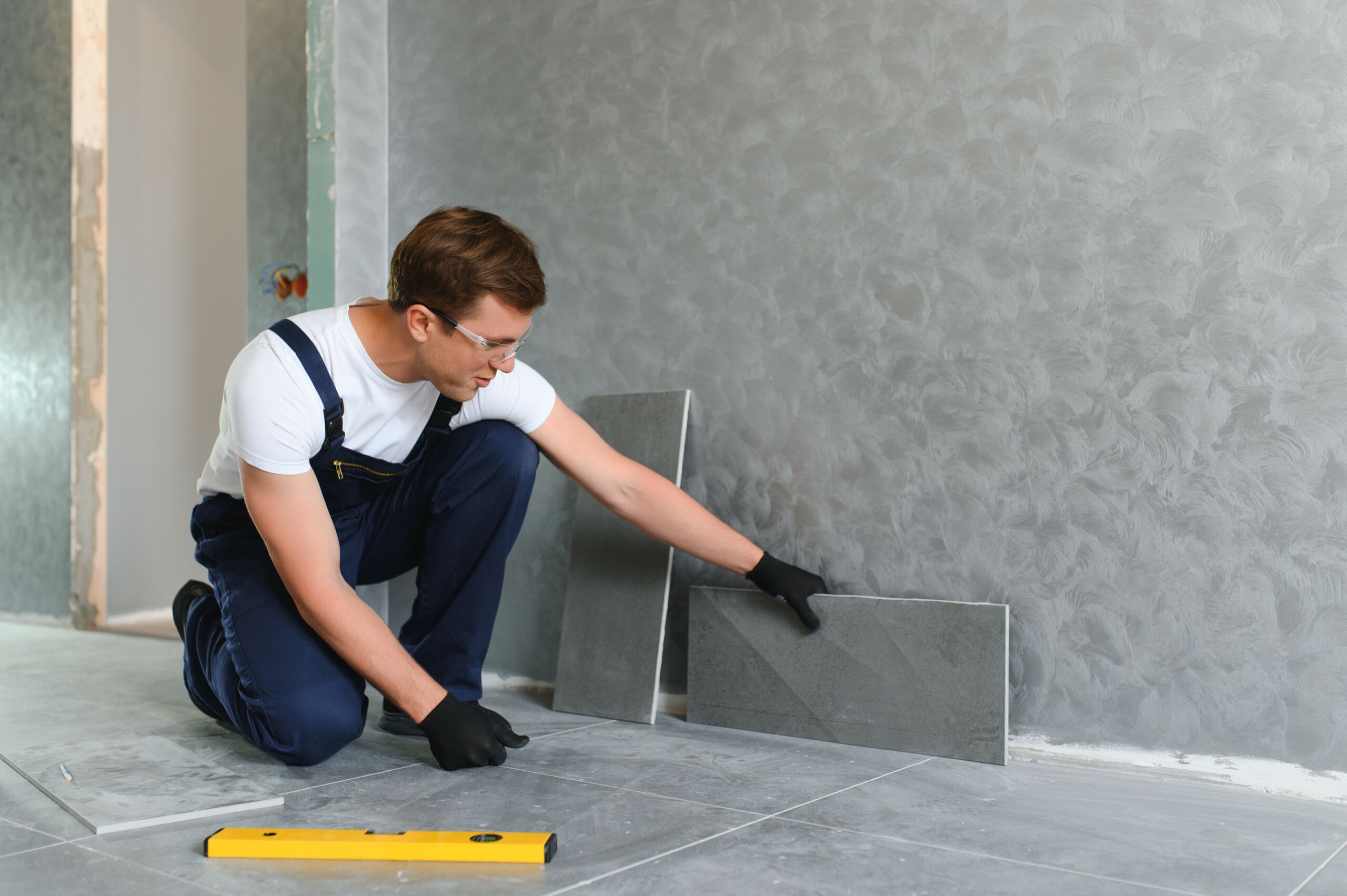 A man wearing gloves and overalls kneels on the floor, positioning large grey tiles against a wall, with a spirit level placed nearby.
