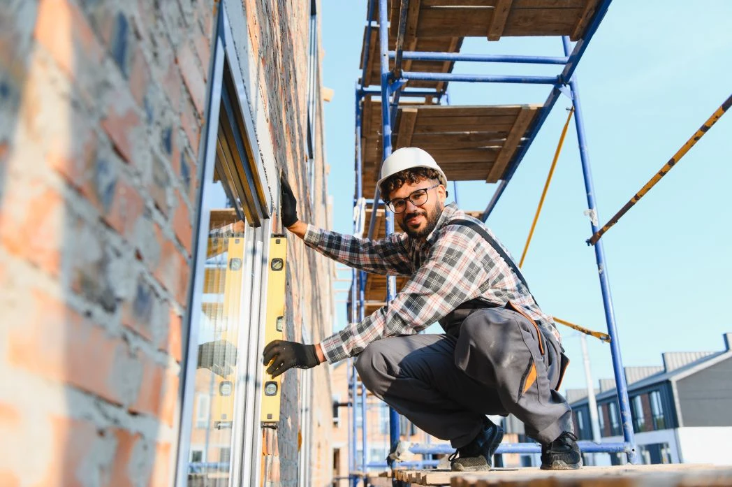 Construction worker in a hard hat uses a spirit level against a window frame on a brick building, with scaffolding and buildings in the background.