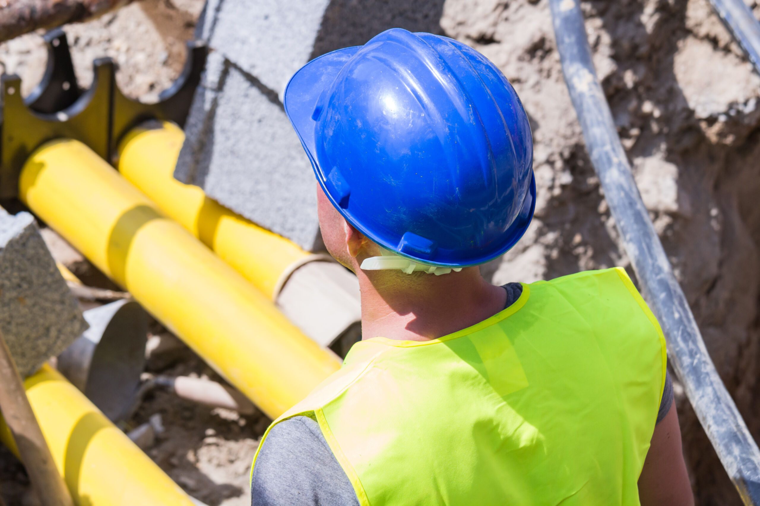 Person in a blue hard hat and yellow safety vest stands near exposed yellow pipes and cables at a building site.