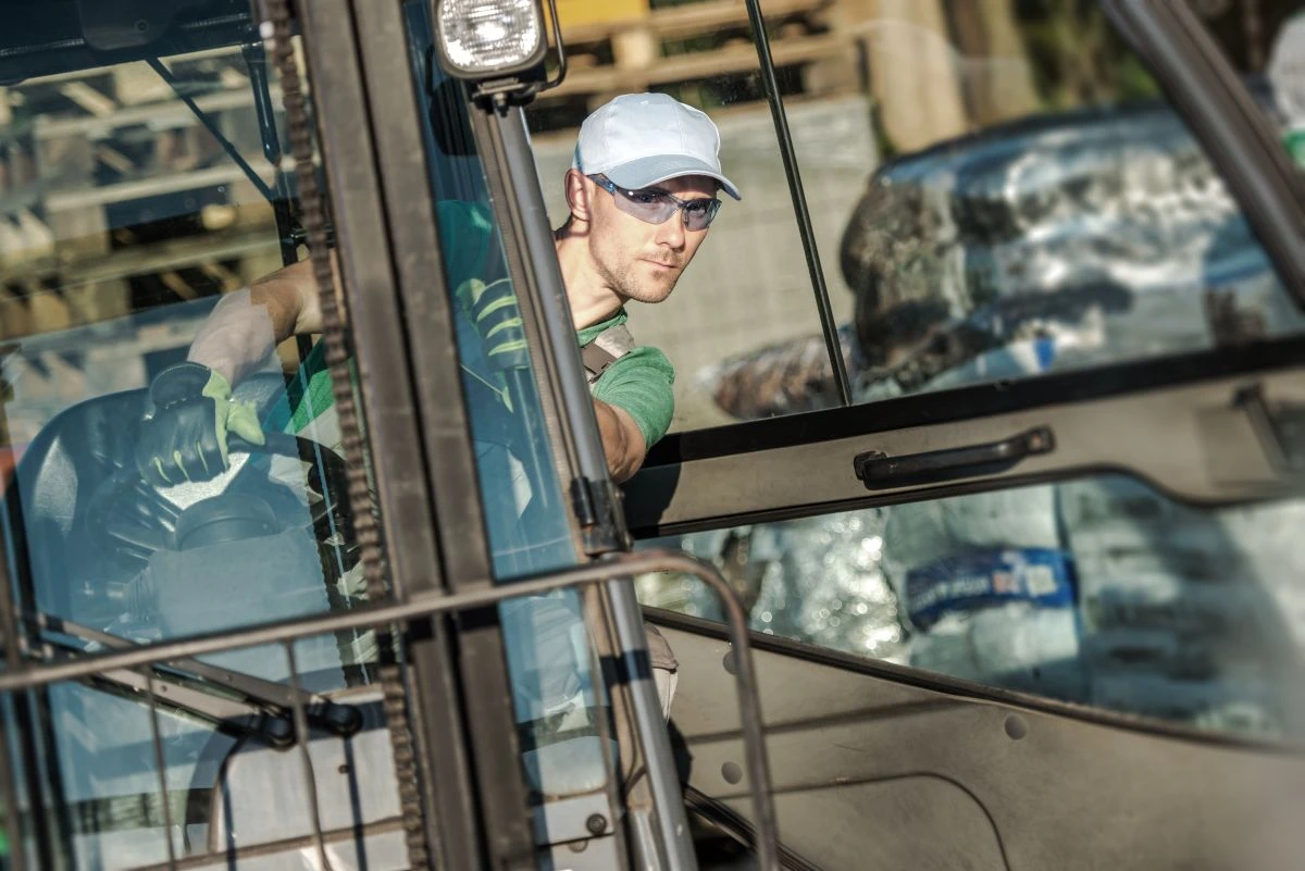 A man wearing a white cap and sunglasses operates a forklift, turning to look behind him whilst inside the cab.