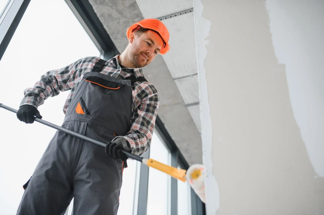 Man in a checked shirt, dungarees, and orange hard hat painting a white interior wall with a roller on an extension pole.