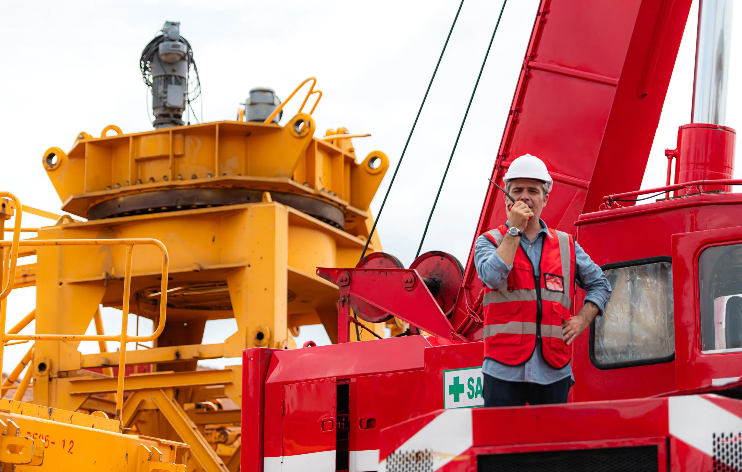 A construction worker in a red safety vest and white helmet speaks into a walkie-talkie while standing on red machinery near large yellow industrial equipment.