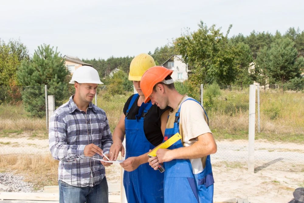 Three men, two in blue overalls and hard hats, and one in a checked shirt and white hard hat, review papers outdoors at a building site.