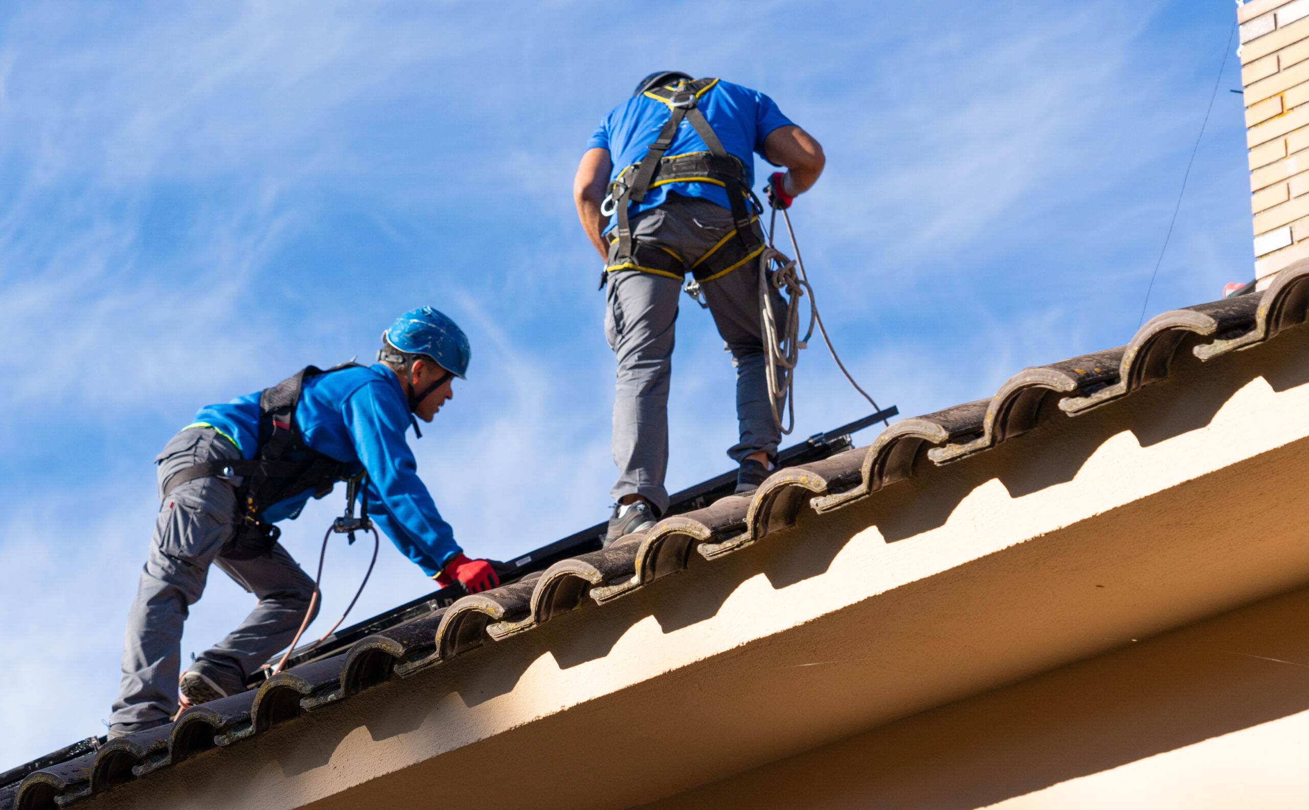 Two workers wearing safety gear and harnesses are on a tiled roof under a blue sky, carrying out maintenance or repairs.