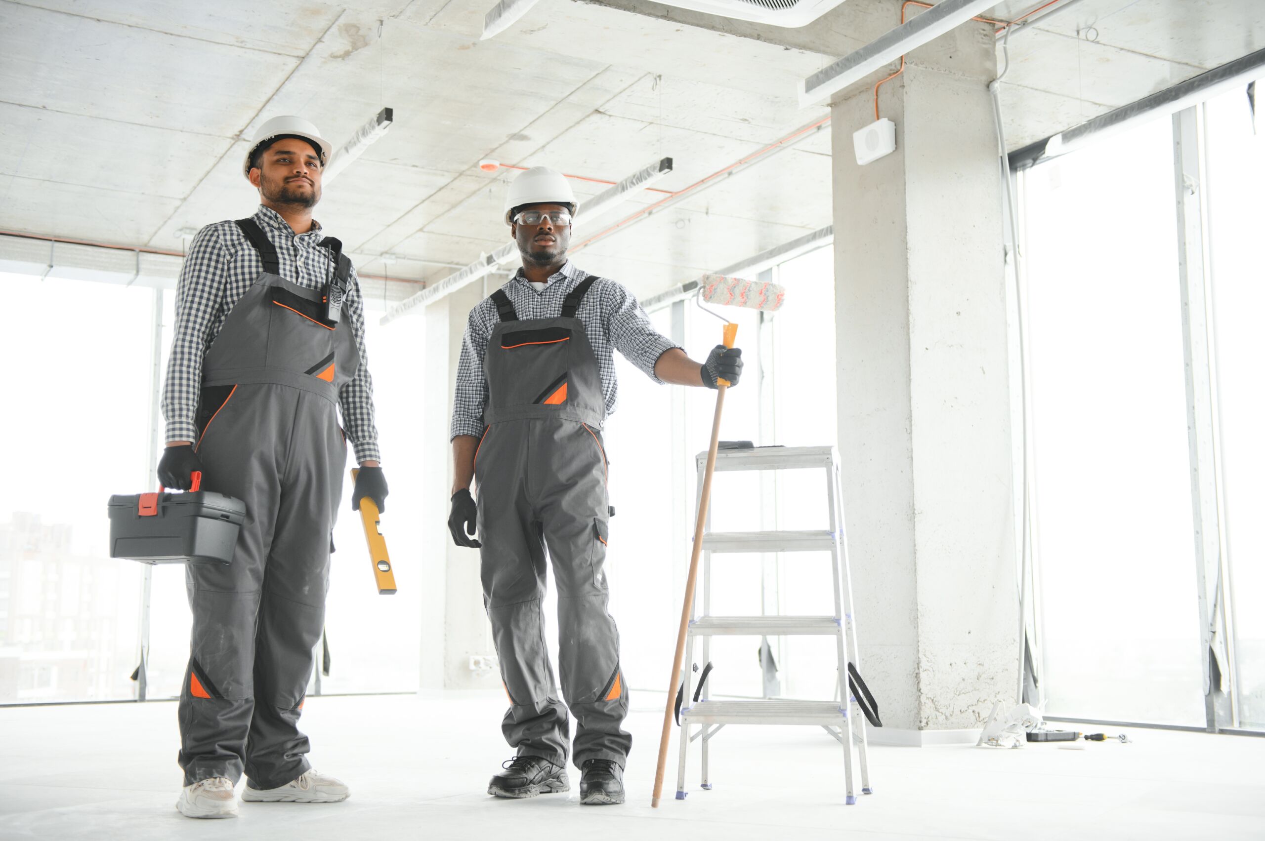 Two construction workers in uniforms stand in a bright, unfinished room with a toolbox, spirit level, paint roller, and stepladder.