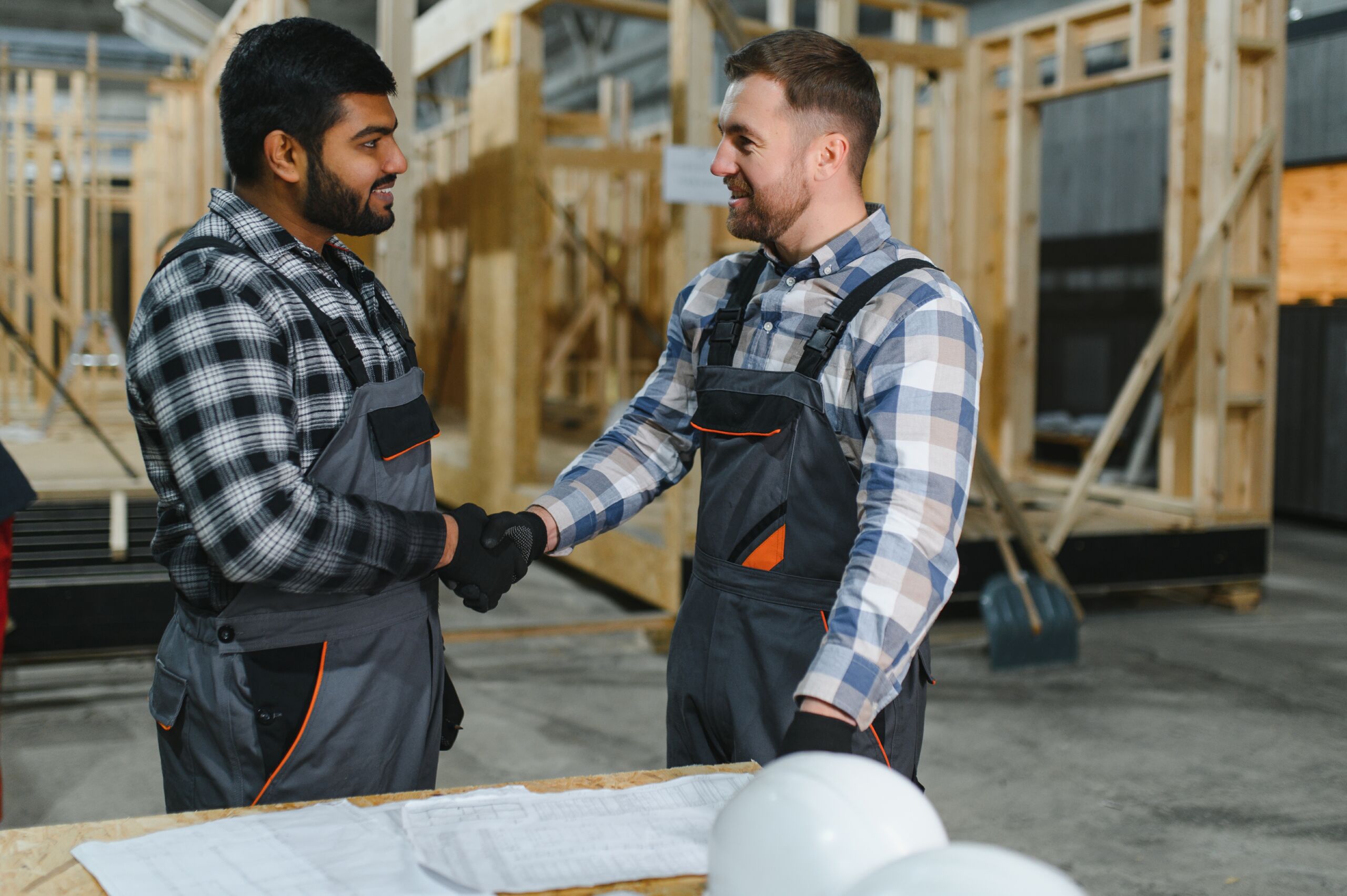 Two construction workers in checked shirts and overalls shake hands in a workshop, standing by plans and hard hats, unfinished wooden frames in the background.