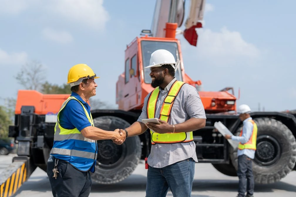 Two construction workers in safety vests and helmets shake hands at a building site with heavy machinery in the background.