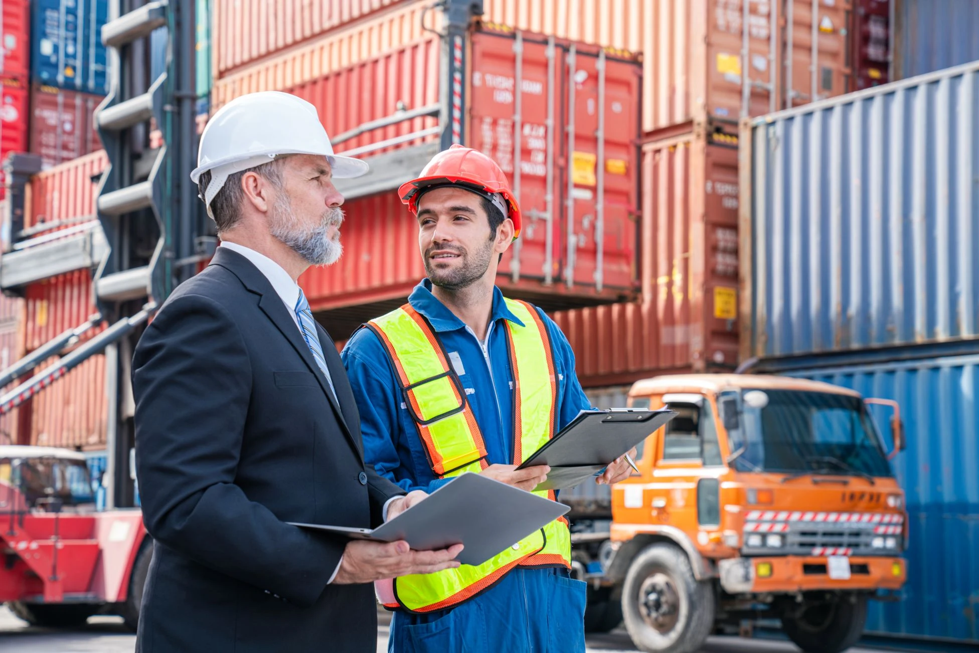 Two men wearing safety helmets and holding clipboards stand in a shipping yard surrounded by stacked cargo containers and a lorry.