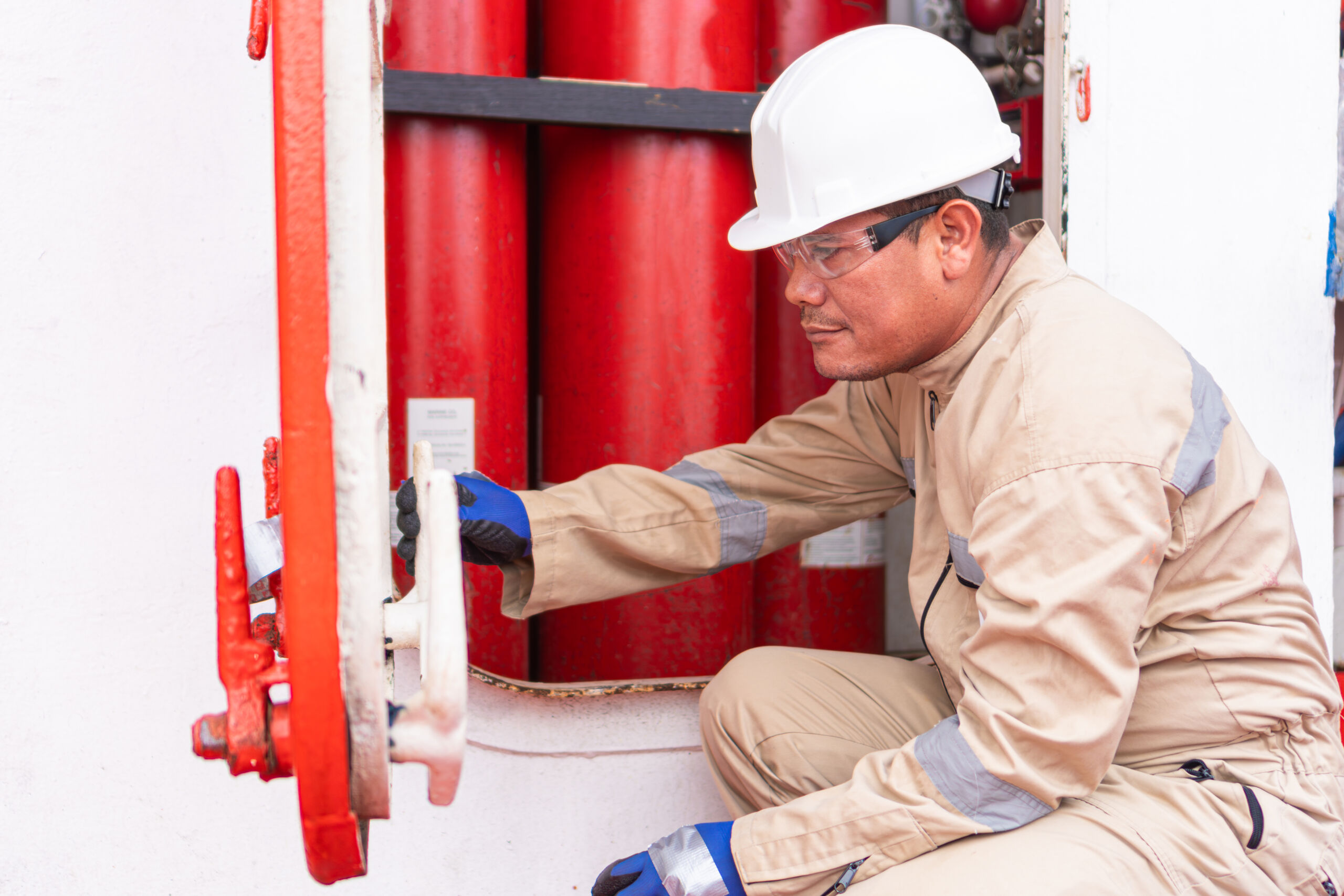 A worker in a hard hat and gloves operates a red valve near large red cylinders, possibly handling safety or maintenance tasks.