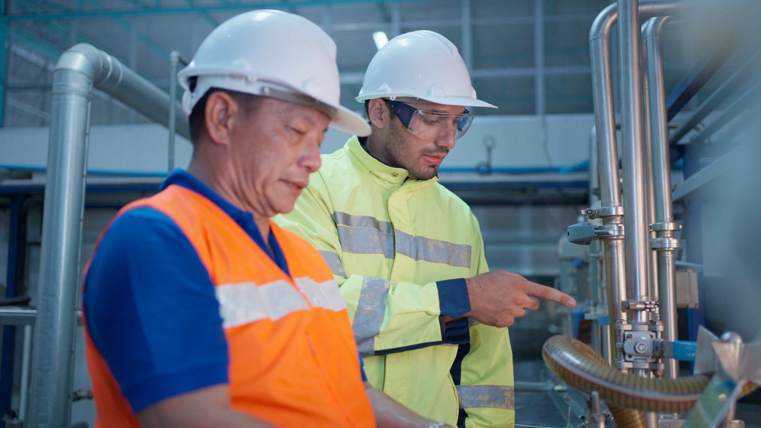 Two workers wearing hard hats and safety vests inspect industrial equipment in a factory setting. One is adjusting controls whilst the other points at a section of the machinery.