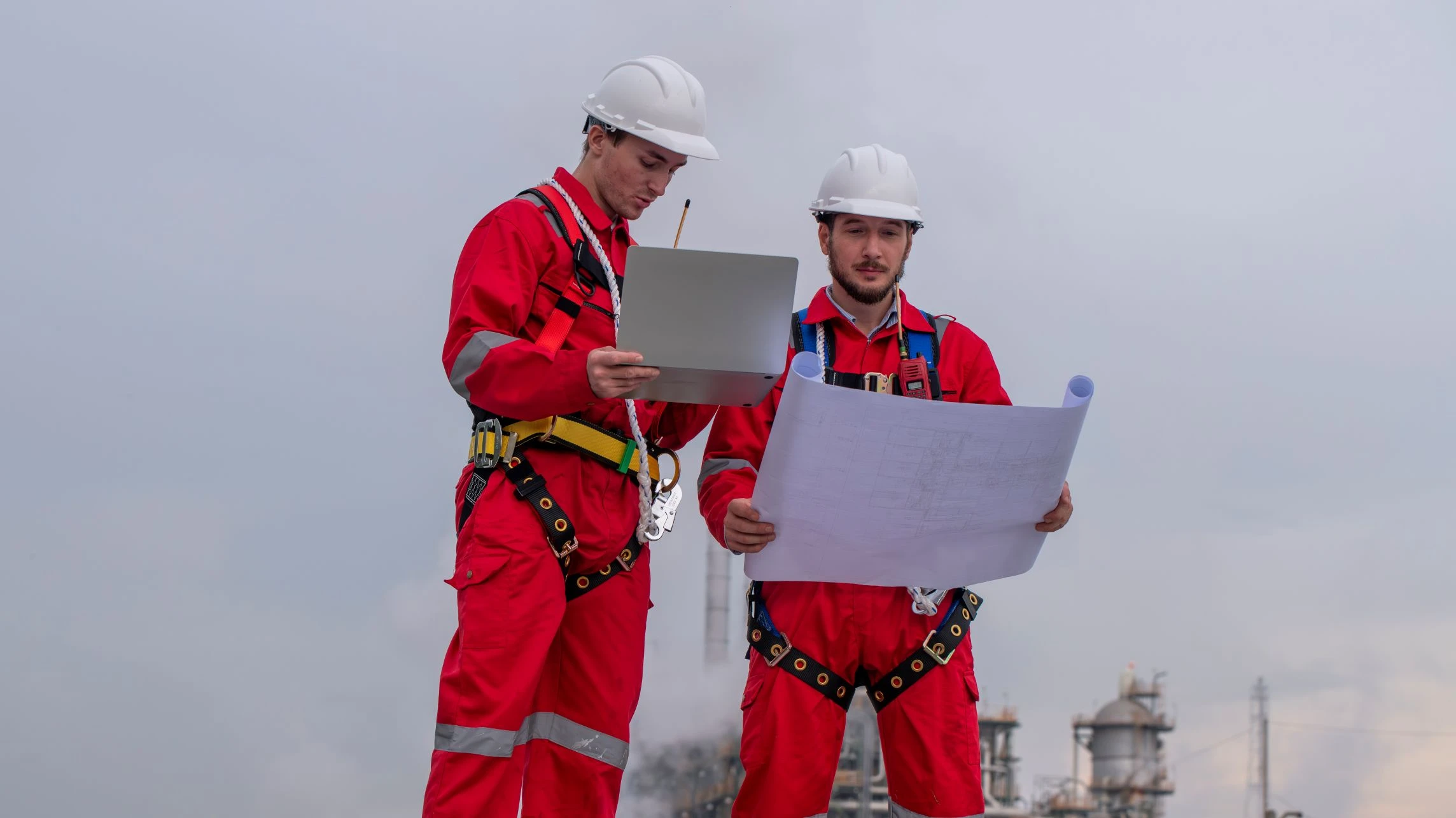 Two workers in red protective suits and white helmets review documents and blueprints outdoors at an industrial site.