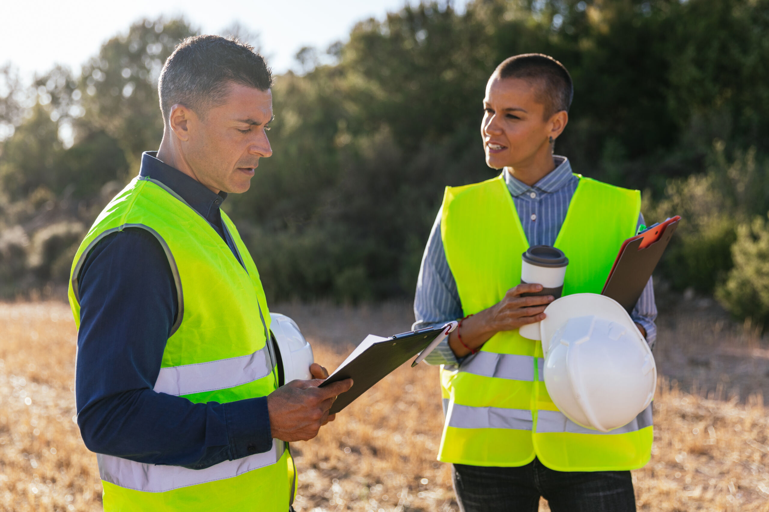 Two people wearing yellow safety vests stand outdoors, holding clipboards and a hard hat, discussing work; trees and dry grass are visible in the background.