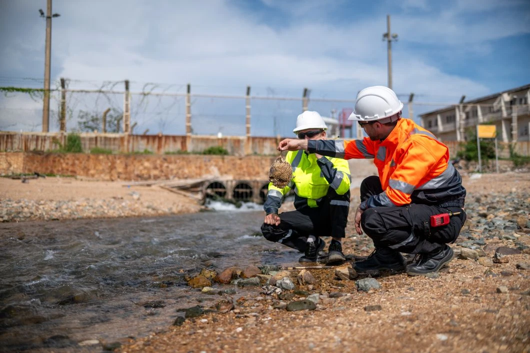 Two workers in safety gear kneel by a stream, collecting a water sample near a drainage outlet in an outdoor, industrial area.