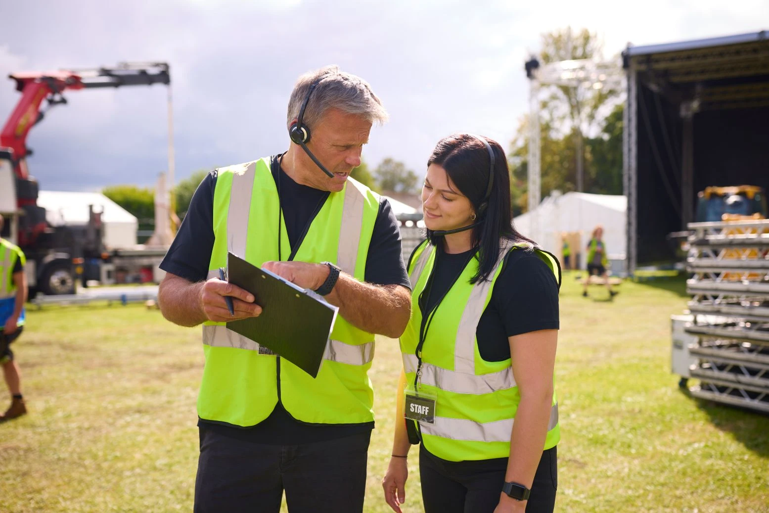 Two event staff in yellow safety vests and headsets discuss a clipboard outdoors near a stage and equipment during daylight.