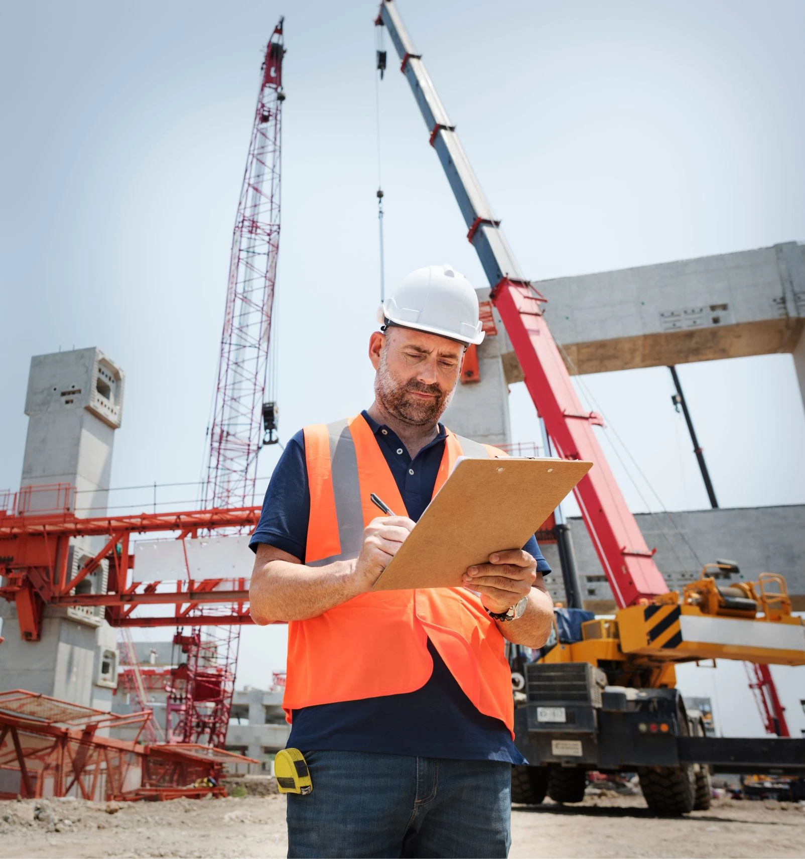 A man in a hard hat and orange safety vest writes on a clipboard at a construction site with cranes and concrete structures in the background.