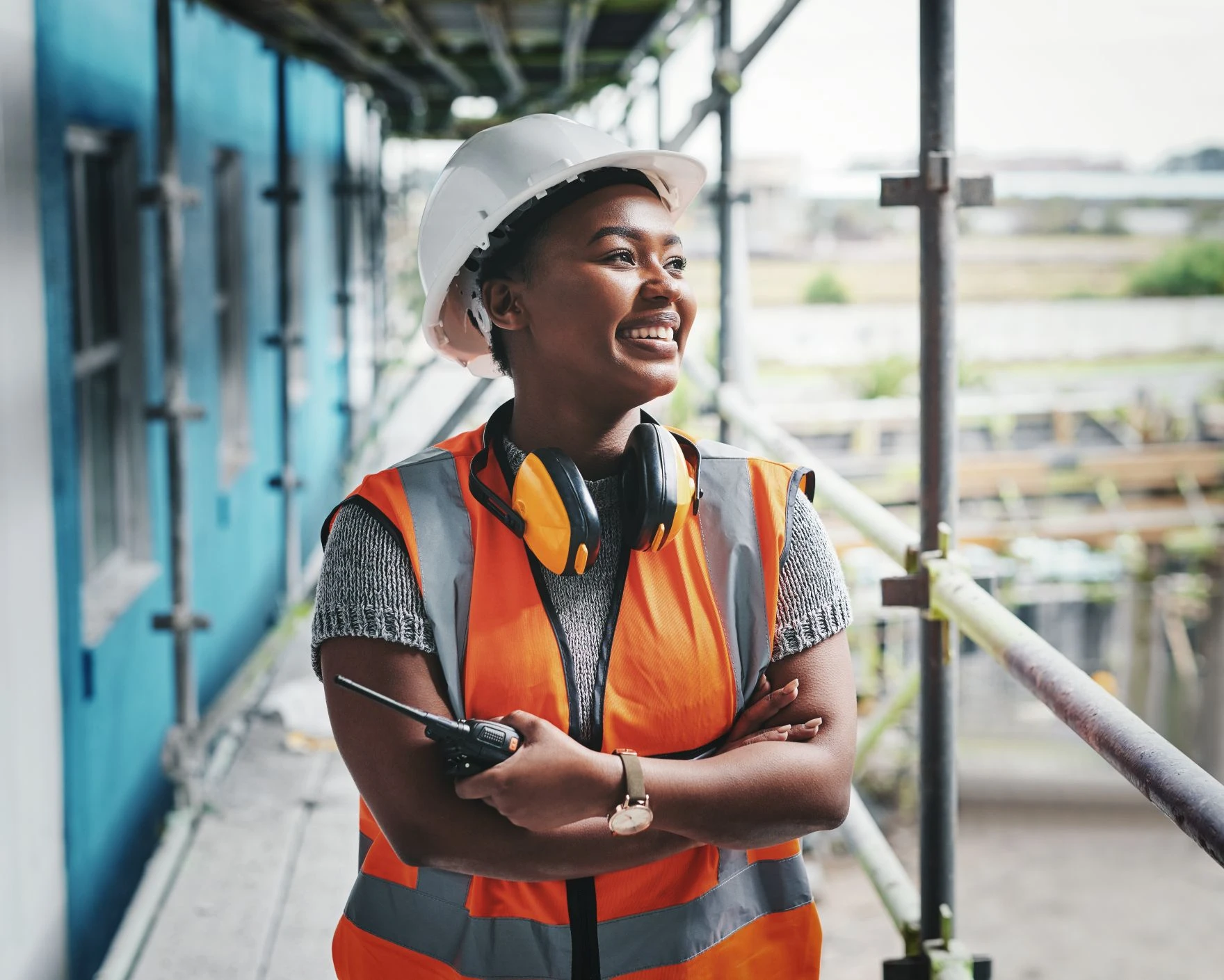 Person in an orange safety vest and white hard hat stands on a building site, holding a walkie-talkie and smiling, with protective headphones around their neck.