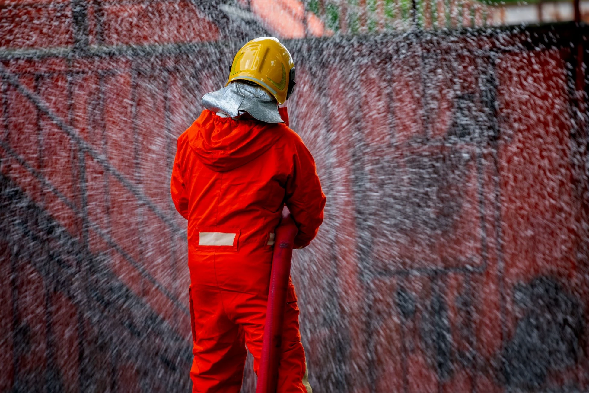A firefighter in orange gear and a yellow helmet sprays water from a hose, creating a mist in front of a red wall.