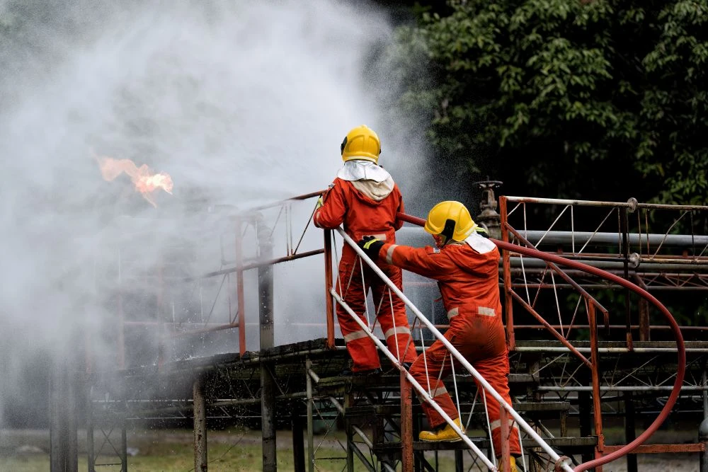 Two firefighters in orange suits and yellow helmets spray water onto flames from a metal structure outdoors, with trees in the background.