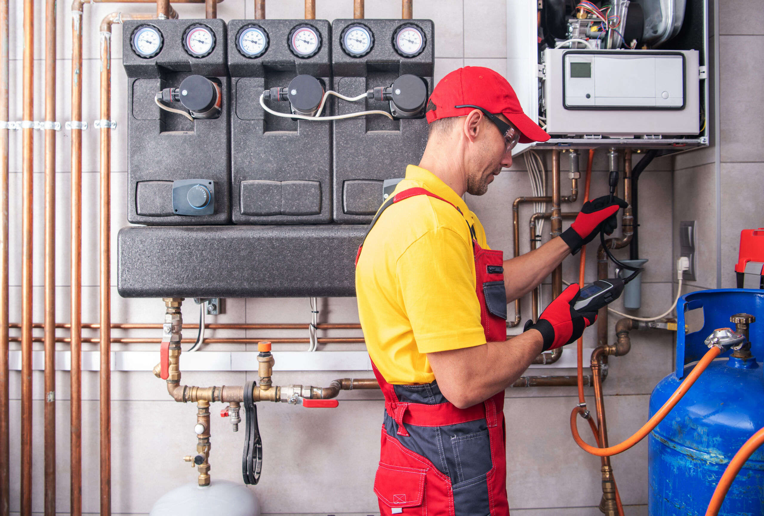A technician in red overalls and a cap checks gauges and controls on a heating system with exposed copper pipes and a blue gas cylinder.