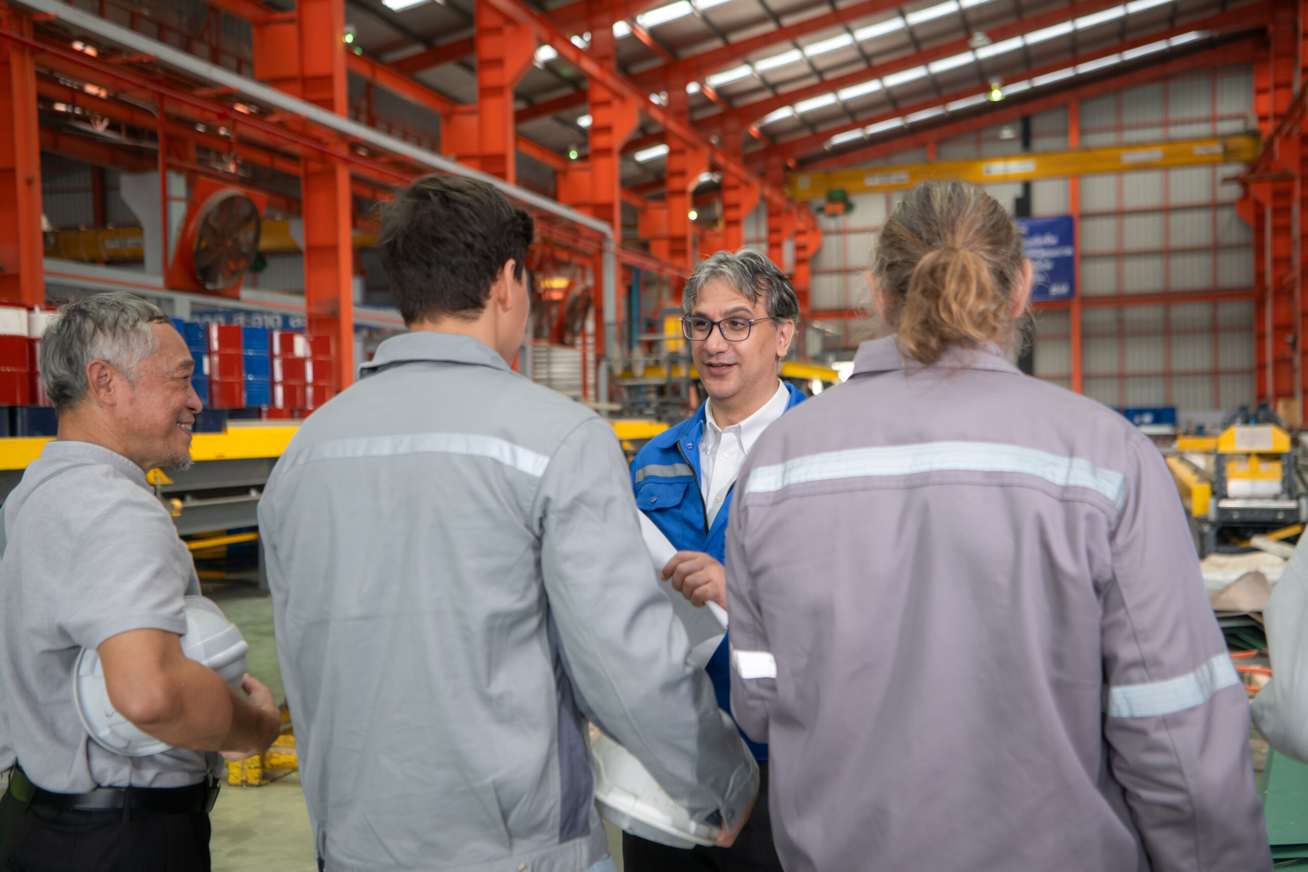 Four people stand talking in an industrial warehouse with red beams, safety gear, and machinery visible in the background.