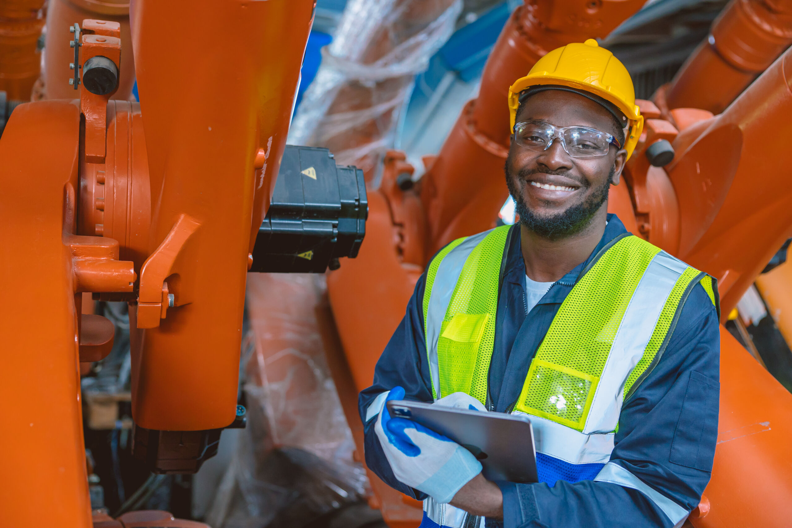 Smiling worker in safety gear holds clipboard, standing among large orange industrial machines in a factory setting.