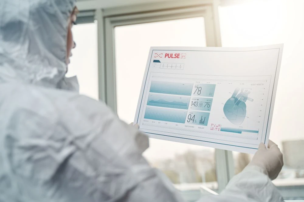 Person in protective suit holding a transparent screen displaying vital signs, heart rate, and medical charts in a bright clinical setting.