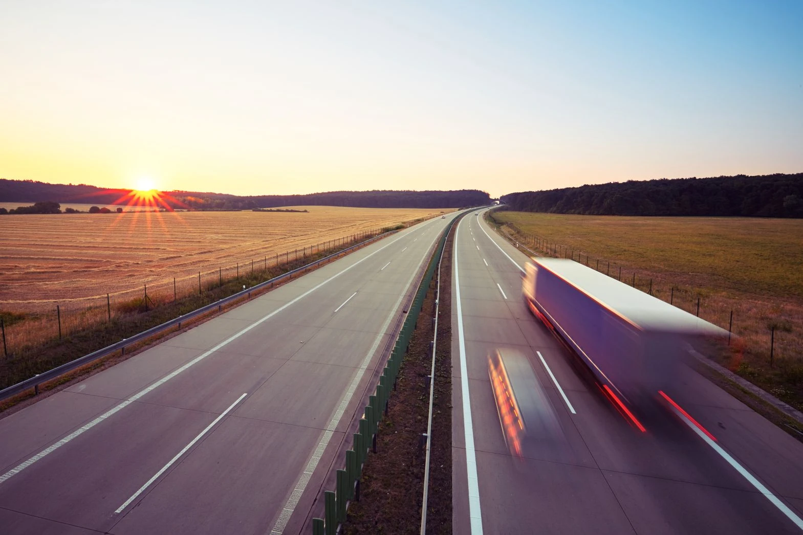 A blurred lorry drives on an empty motorway through open fields at sunset, with clear skies and distant trees in the background.