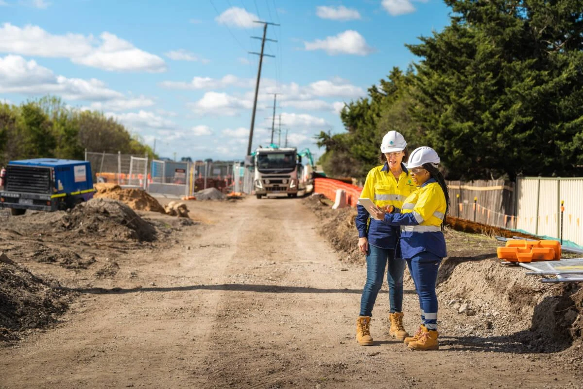Two construction workers in safety gear stand on a dirt road at a construction site, reviewing plans. Equipment, fencing, and trees are visible in the background under a partly cloudy sky.