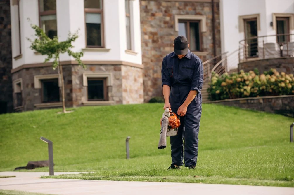 A person in dark work clothes uses a leaf blower on a lawn in front of a building with stone walls and large windows.