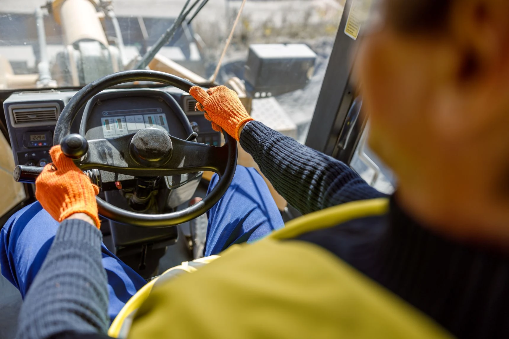 Person wearing orange gloves and a high-visibility vest steering heavy machinery, viewed from behind inside the vehicle cab.