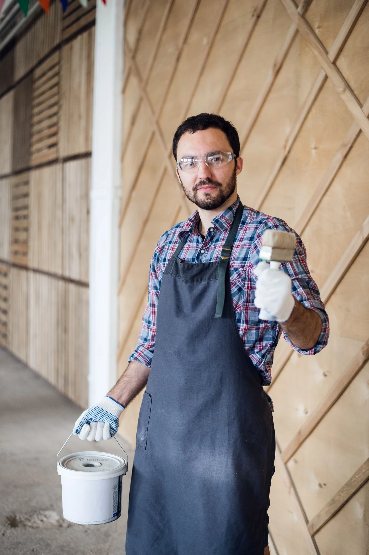 A person wearing gloves, safety glasses, an apron, and a checked shirt holds a paint roller in one hand and a paint tin in the other, standing in front of a wooden wall.