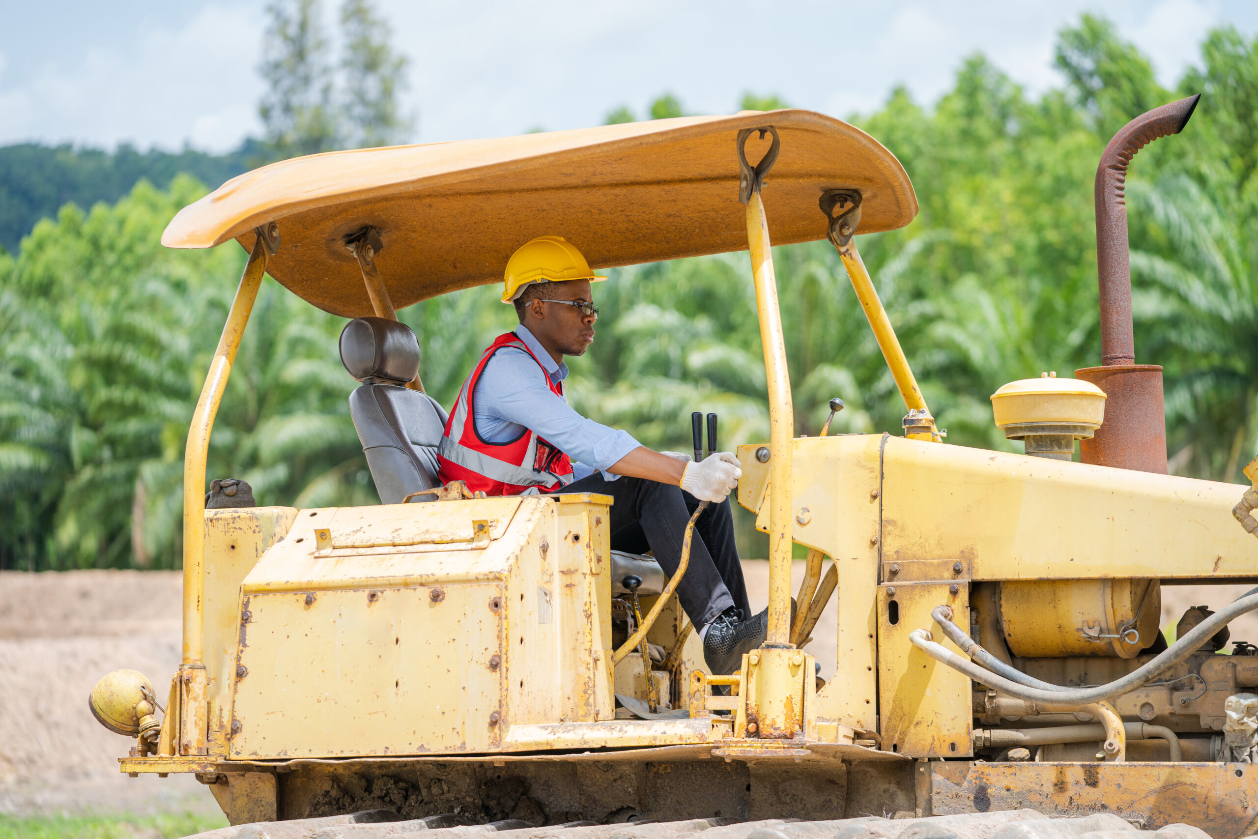 A person wearing a yellow hard hat and red safety vest operates a yellow bulldozer at a construction site with trees in the background.